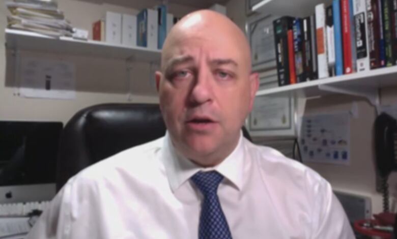 A bald man, wearing a white shirt and a blue tie, sits in front of bookshelves in an office.