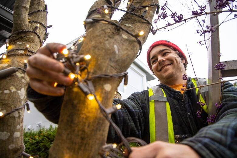 A man smiles as he wraps Christmas lights around an exterior branch.