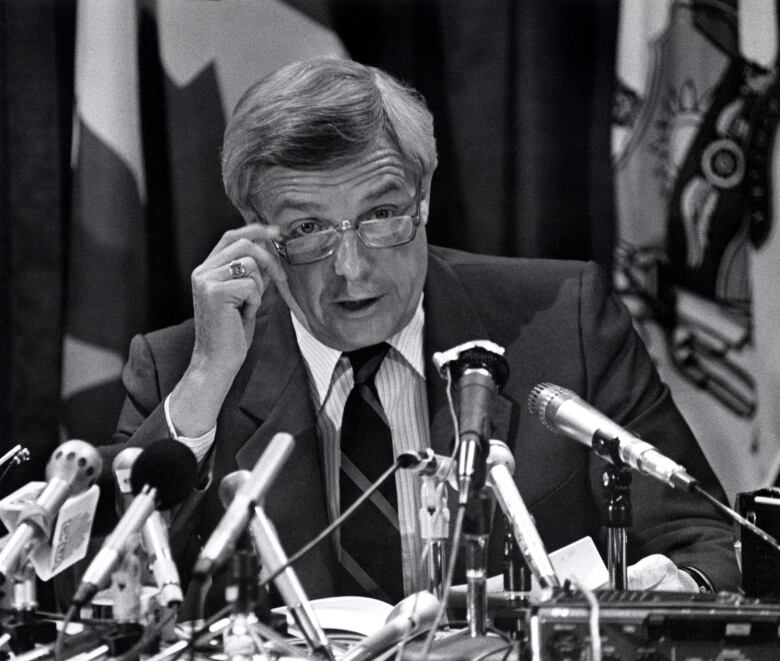 A politician lowers his glasses as he sits behind several microphones, in a black-and-white photo of former Alberta premier Peter Lougheed from 1984.