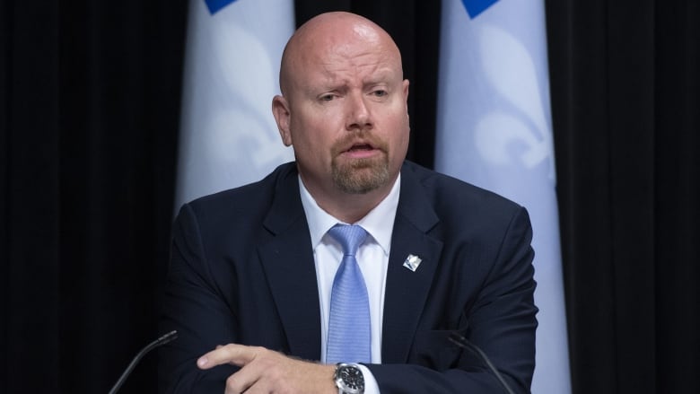 Ian Lafrenire speaking at a conference flanked by Quebec flags. 