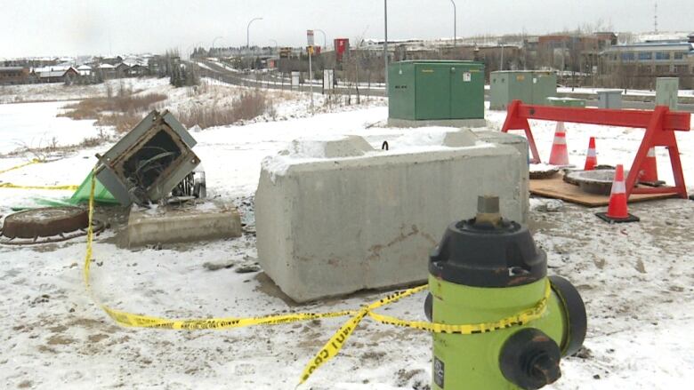 a yellow fire hydrant is pictured with police do-not-cross tape wrapped around it in the foreground, with a large cement block and toppled electrical box farther afield on a snowy ground next to a roadway.