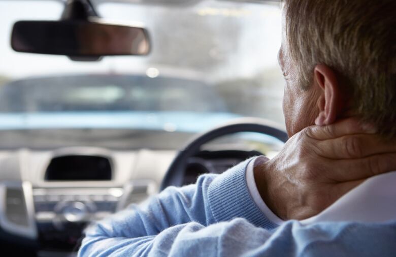 A man sits in the driver's seat of a vehicle with his hand holding his neck.