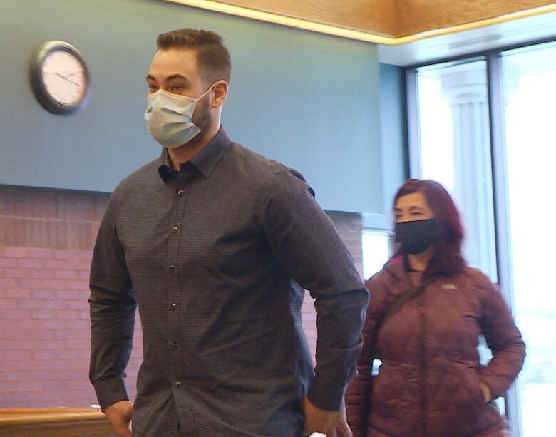 A tall young man wearing a dark collared shirt and face mask walks in front of a woman wearing a red coat.