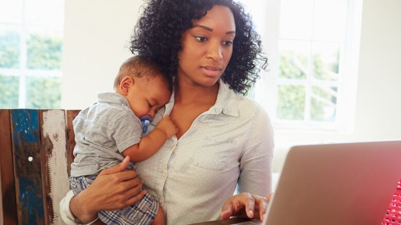 A woman holds a child while sitting at a desk and working at a laptop computer. 