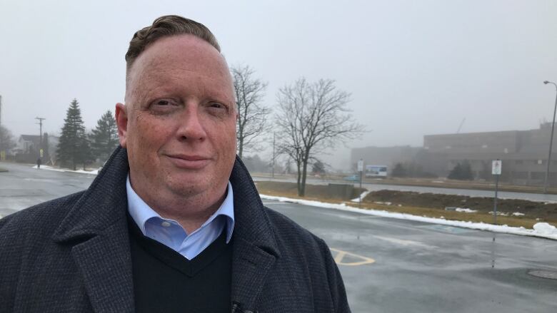 Man in dress shirt and jacket standing outdoors with parking lot behind him.