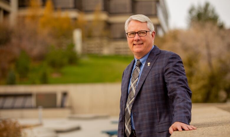 Mike Mahon, a grey-haired man with classes in a business suit, stands in front of a college building surrounded by trees.