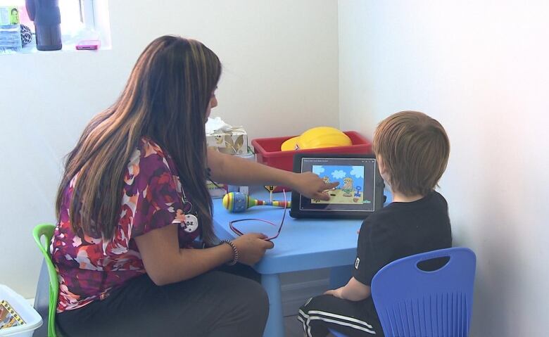 A woman and a boy seated at a small blue plastic table look at a tablet.