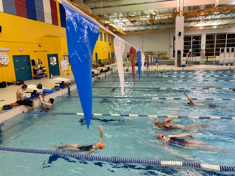 Large indoor swimming pool with designated swimming lanes. Some people are swimming, others sit on the side of the pool.