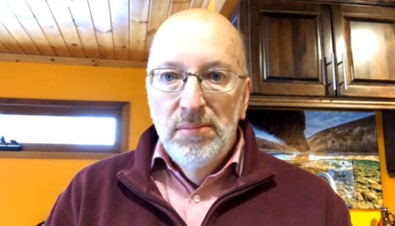 A bald man wearing a burgundy sweater sits in a room with yellow walls.