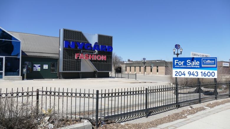 The photo shows a Nygard Fashion store on Broadway in Winnipeg. The building is surrounded by a black metal fence and there are no cars in the parking lot. There is a for sale sign in front of the building.