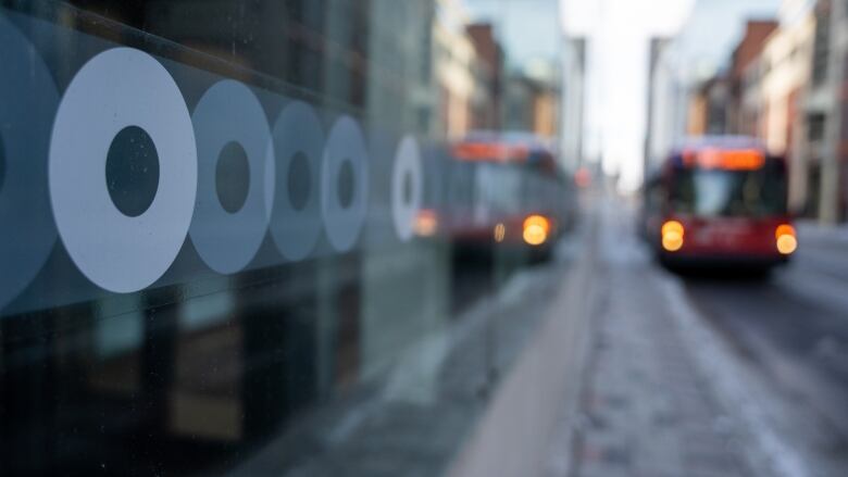 A close-up of a transit agency's logo on the glass wall of a transit shelter, as a bus approaches in the distance.