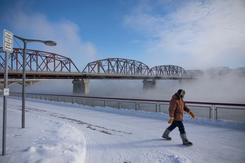 A person in a parka walks on a city path in very cold winter weather as fog rises from a river in the background. The fog partially obscures a bridge.