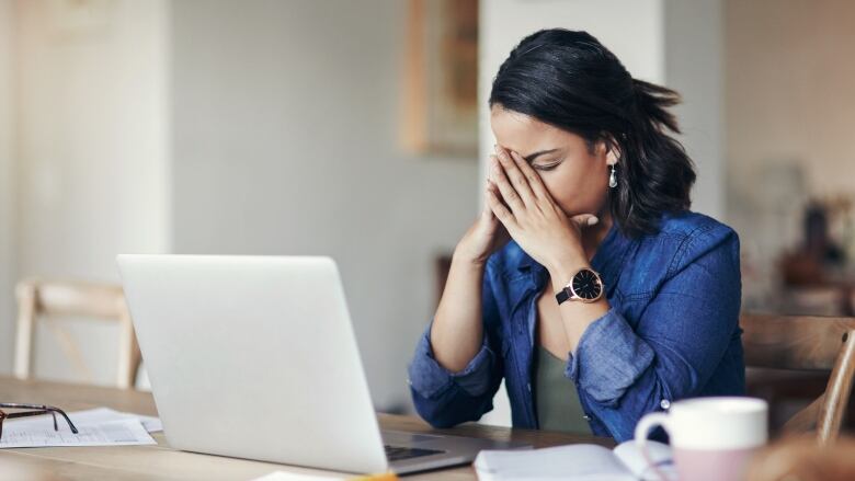 A woman sits at a desk in front of a laptop and holds her face in her hands.