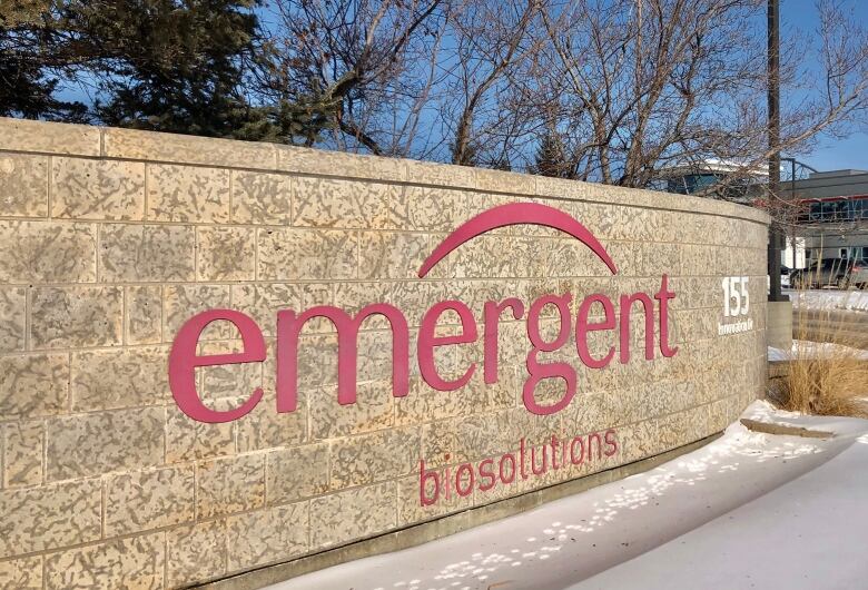 A sign made of rock has the words 'emergent biosolutions' written in red with an arc above it and snow on the ground in front.