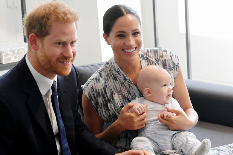  A man with red hair and dressed in a suit next to a woman with long dark hair holding a baby on her lap. 