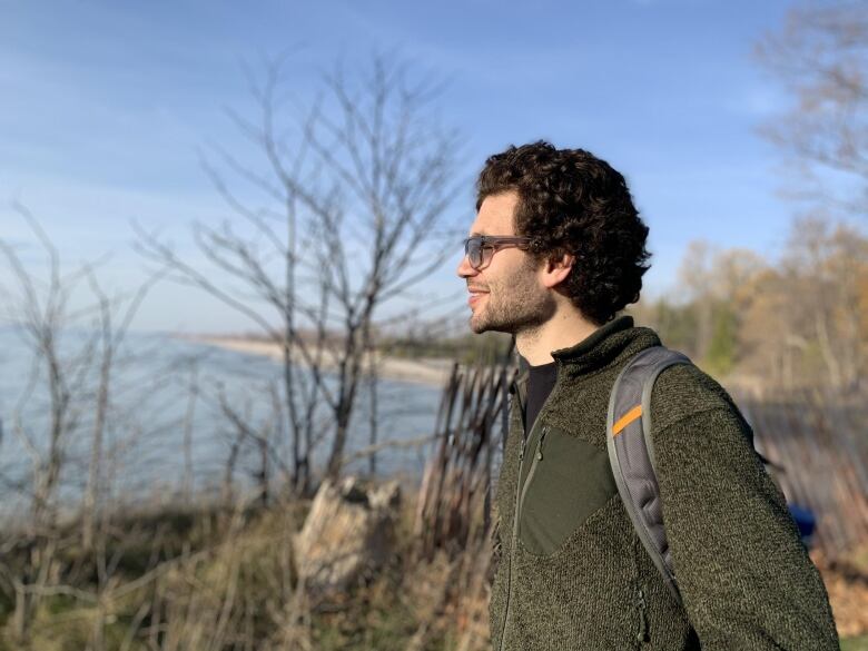 A man wearing a light jacket and backpack stands outdoors under a blue sky, taking in the scenery in the distance.