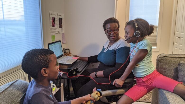 A woman sits at a desk in front of a laptop. A child beside her wearing headphones. A second child is beside them.