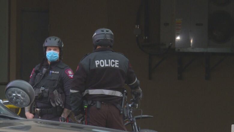 Two police officers in tactical gear stand in front of a wall. One is wearing a mask. 