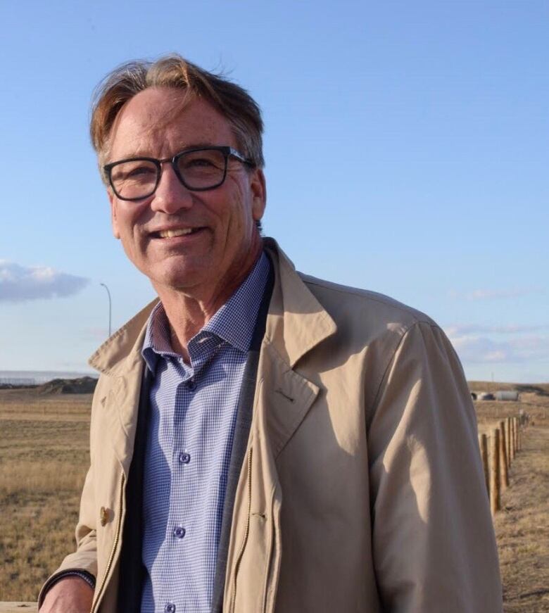 A middle-aged man with dark blond hair and wearing glasses smiles for the camera as he stands outside with a field in the background and a blue sky.