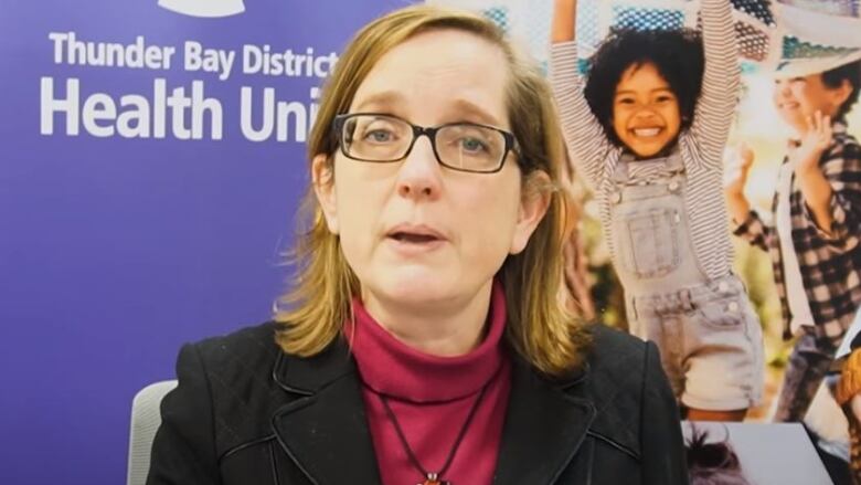 A woman in a black blazer and glasses looks at the camera