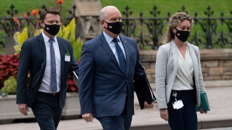 Three people wearing business attire and medical masks cross a street in front of Parliament.