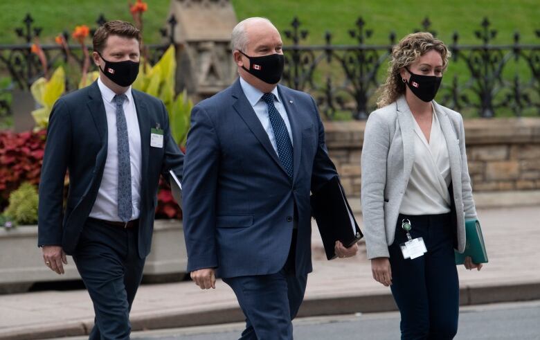 Three people wearing business attire and medical masks cross a street in front of Parliament.