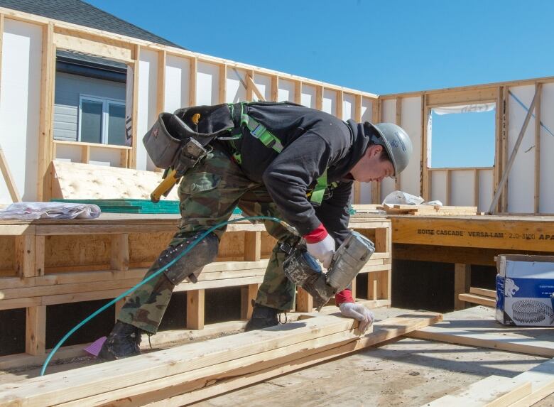 Photo shows a construction worker in a hardhat with a nail gun working on the framework of a single family home.