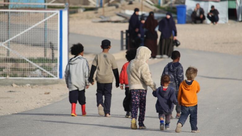 Seven young children, some holding hands, are seen walking along a paved road as women watch in the distance.