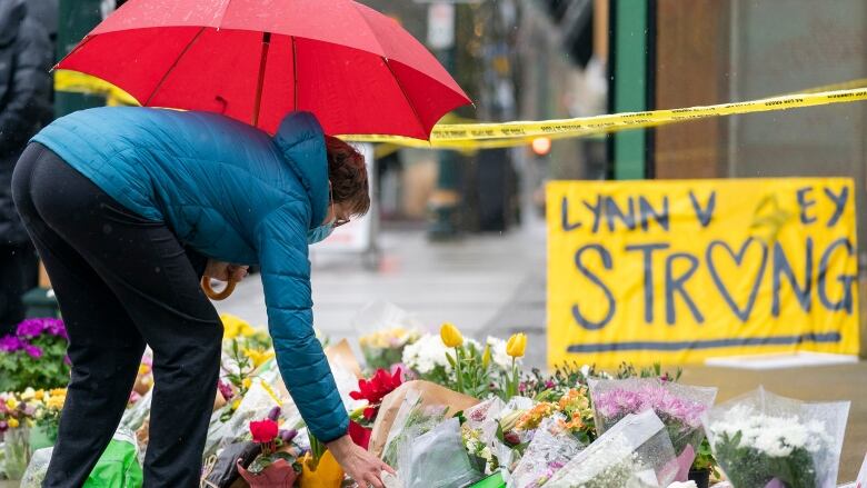 A person in black plants and a blue jacket holds a red umbrella as they place a bouquet of flowers on a pile of tributes on a concrete sidewalk. A yellow sign that says 