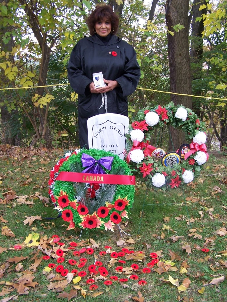 A woman in a black robe stands in front of a grave with flower wreaths, holding a medal. 