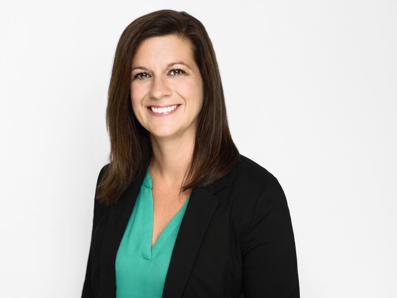 A young woman with medium-length straight dark brown hair stands in front of a while wall. She's wearing a turquoise blouse and dark blazer and smiling at the camera.