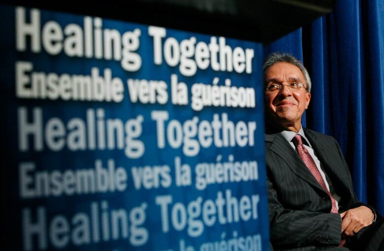 A man with grey hair and glasses sits next to a sign that says 'healing together' in French and English. 