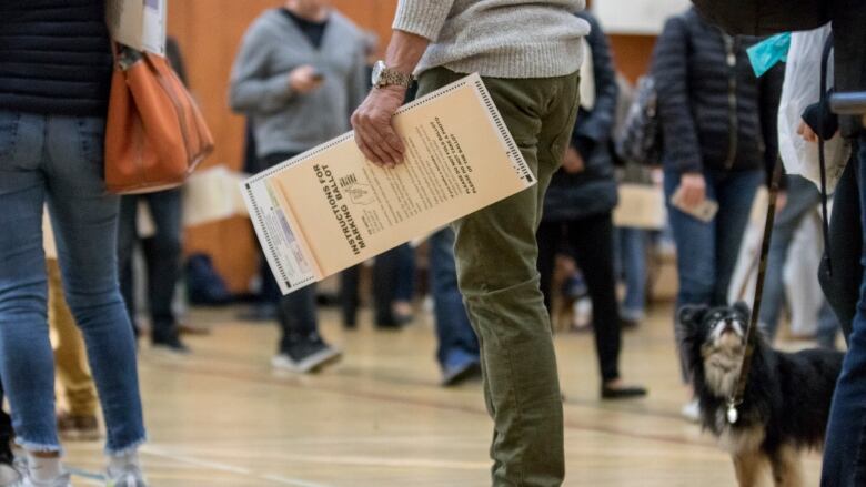 A number of people's legs at a polling station. One person is holding a voter's guide.