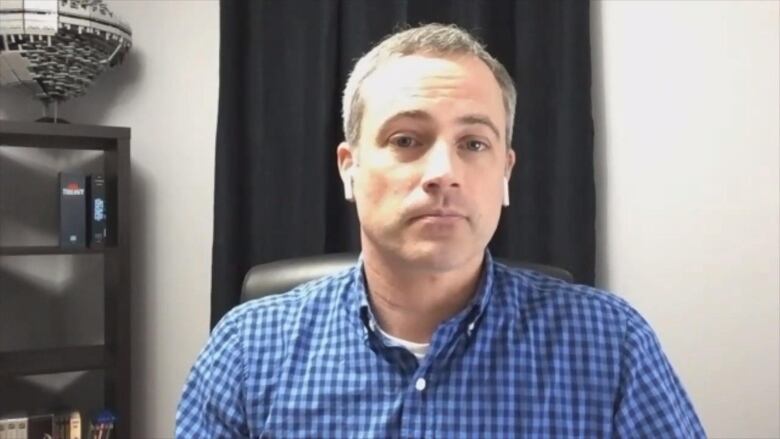 A man with a blue checkered shirt and ear pods sits facing the camera.