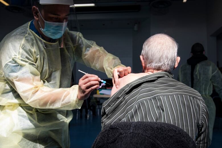 A health-care worker wearing personal protective equipment, including a face shield and mask, administers a vaccine into the arm of an elderly man.