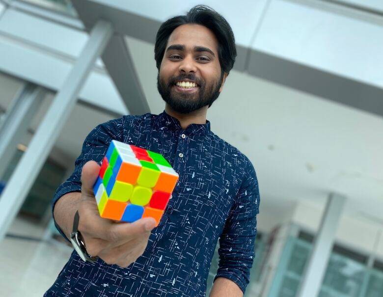 A man smiles at the camera while holding out a Rubik's cube in one hand. 