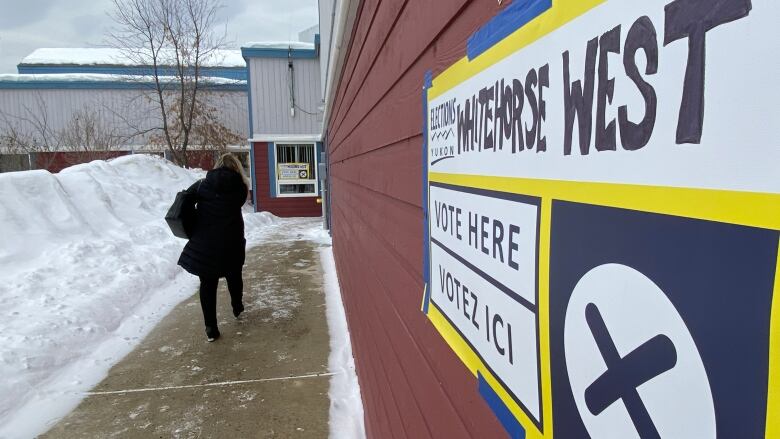 A person is seen from the back walking toward the door of a school building, with a 'Vote Here' sign in the foreground.