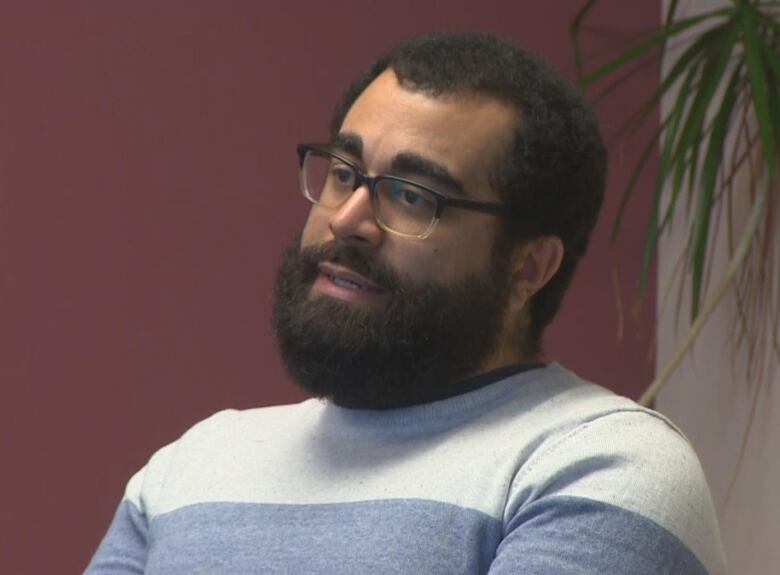 A man with a thick brown beard, brown hair and dark-rimmed glasses wearing a white and blue sweater.