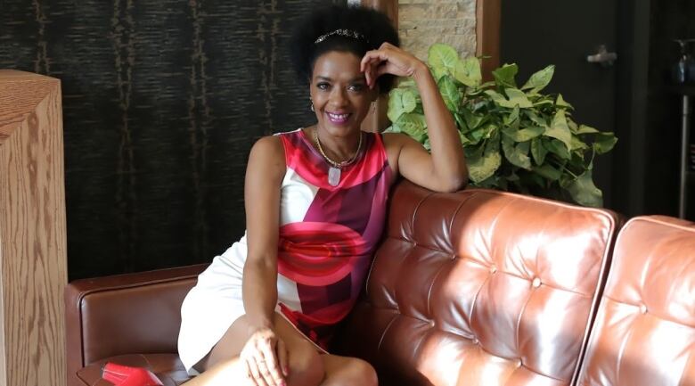 A woman sits sideways on a leather couch, facing the camera and with one elbow propped on the back of the couch.