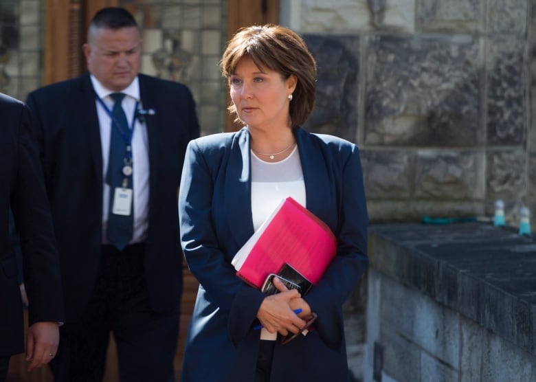 A woman in a blue suit walks outside, attentive, arms-crossed and carrying documents behind a red transparent file cover.