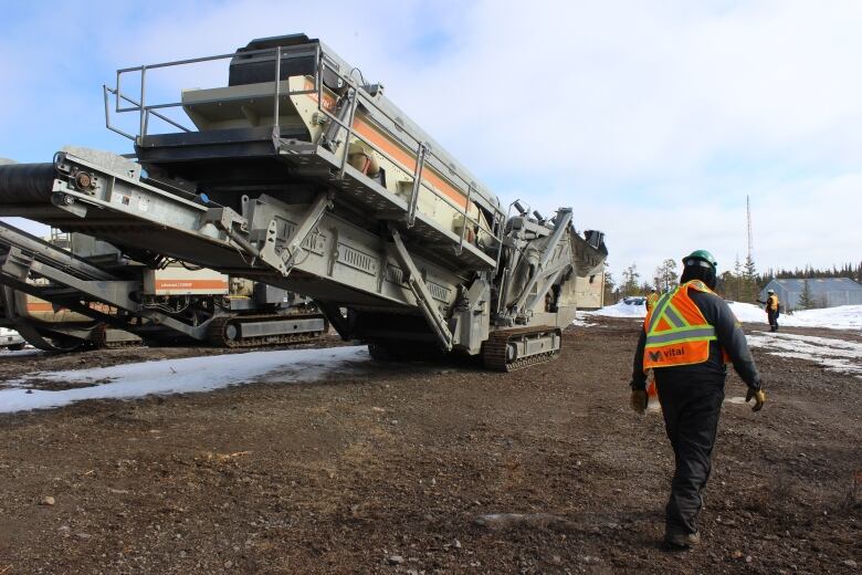 A man in a Vital metals coat walks by a big piece of heavy machinery.