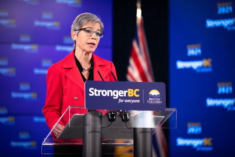 Selina Robinson stands at a podium with 'StrongerBC for Everyone' on it. She is a white woman with blue thin-framed glasses, wearing a red top.