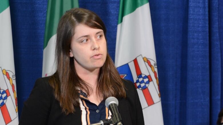 A woman stand at a microphone with flags and a curtain behind her.