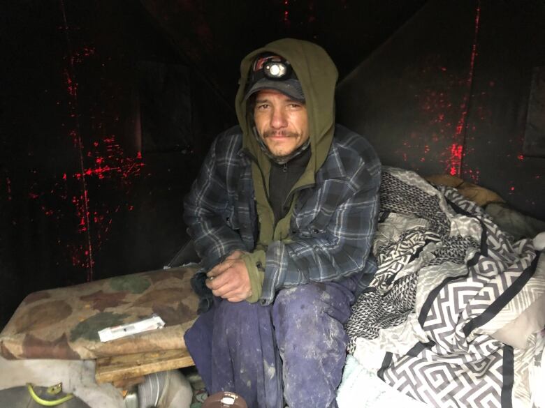 A photo of a man in many layers of clothes and a hood over his head, sitting on an older looking mattress. 