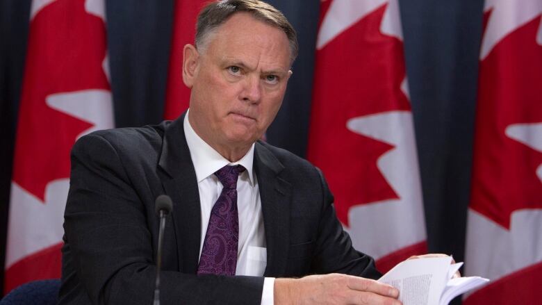 A man in a suit opens a report at a table in front of a row of Canadian flags.