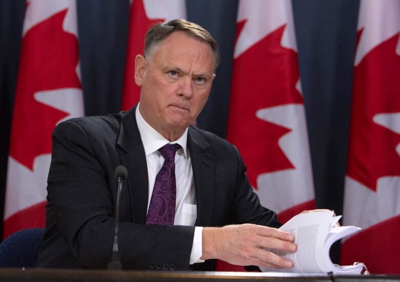 A man in a suit opens a report at a table in front of a row of Canadian flags.