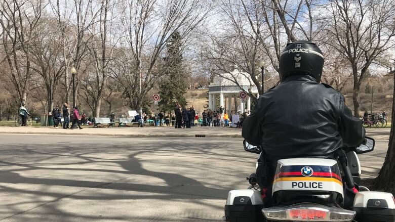 A police officer seated on a police motorcycle watches a large group of people attending an anti-lockdown rally at a sunny park.   