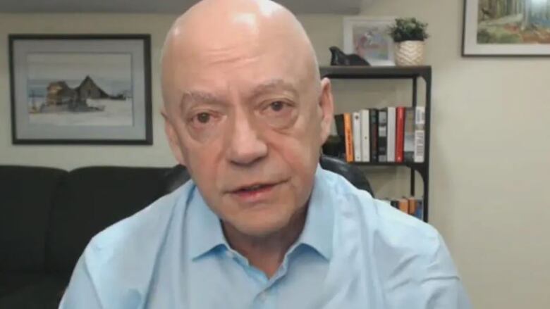 Noel Gibney sits in front of a couch and book case and looks directly at the camera during an interview.