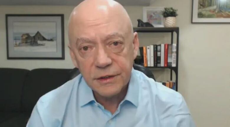 Noel Gibney sits in front of a couch and book case and looks directly at the camera during an interview.