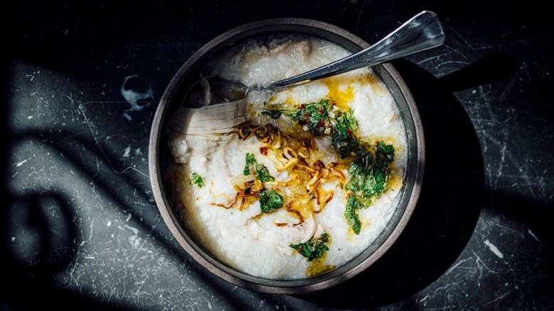 Overhead shot of a grey bowl of kanji, a white, porridge-like soup made with rice and chicken with oil, fried onions and chopped green herbs on top. A spoon sits in the bowl. The bowl is on a black marble table.